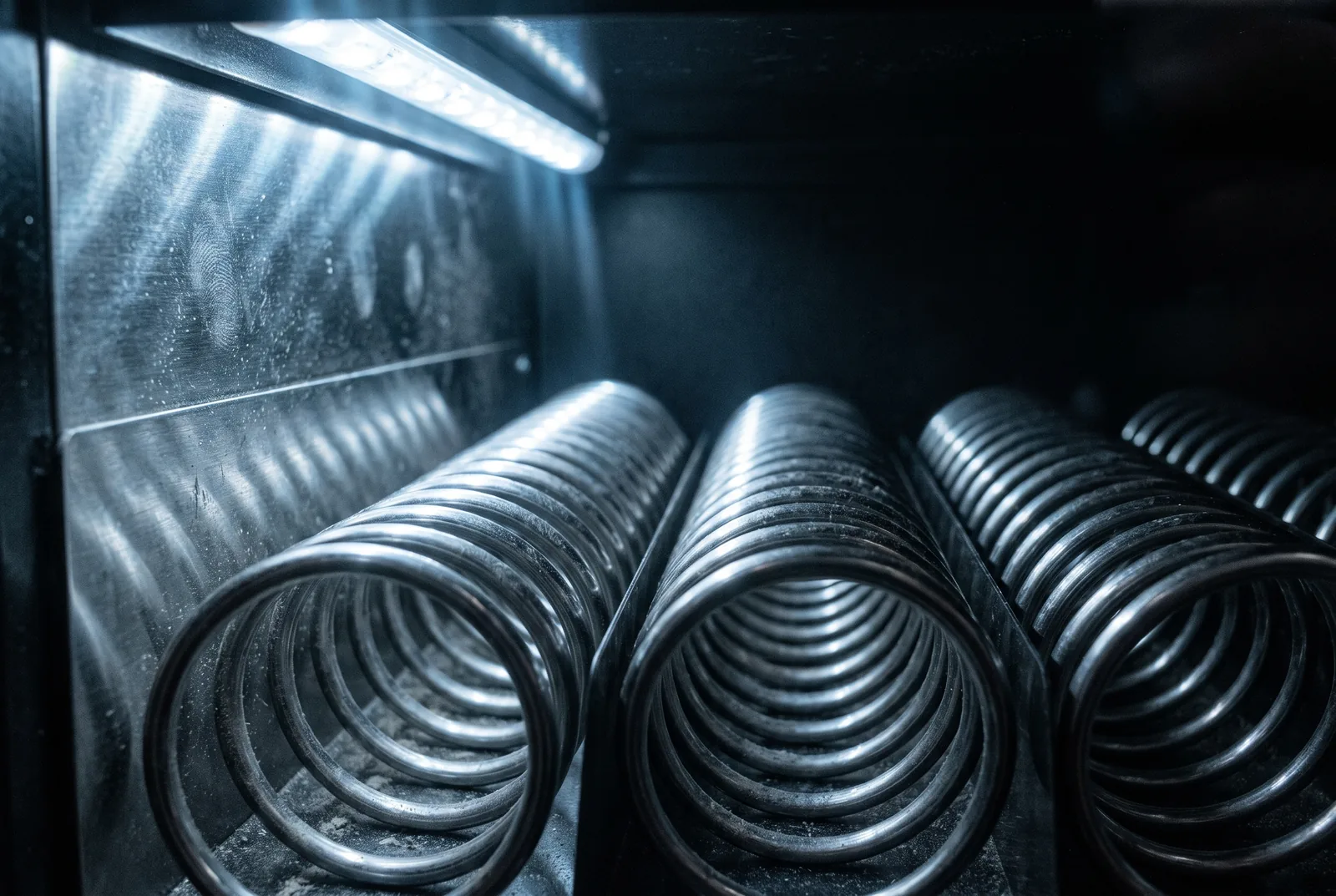 Close detail of chrome dispensing coils inside a retail cabinet, lit by a single LED strip.
