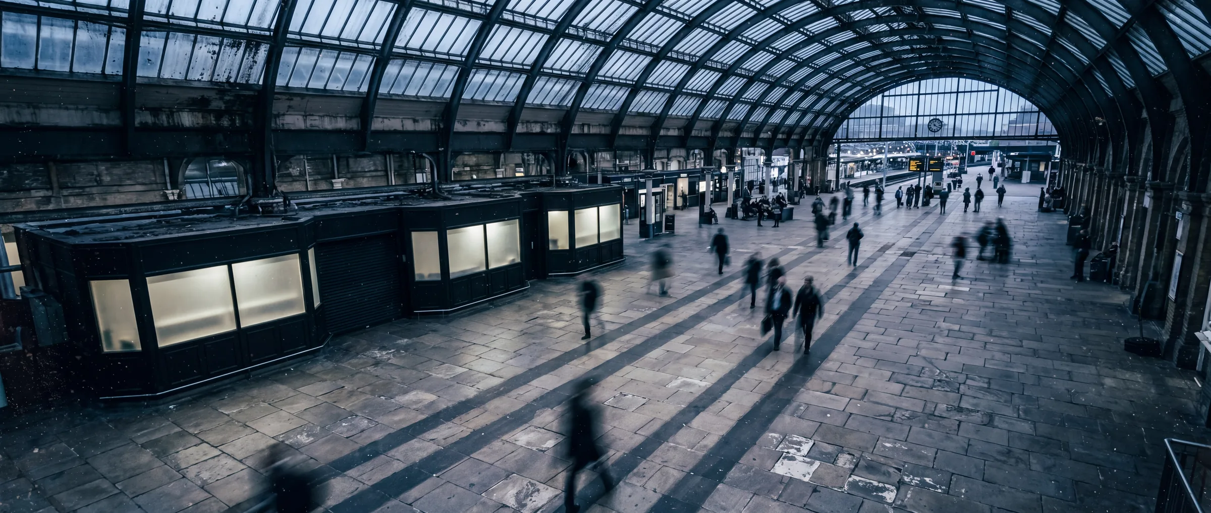 A British railway station concourse in late afternoon, passengers crossing under a vaulted glass roof.
