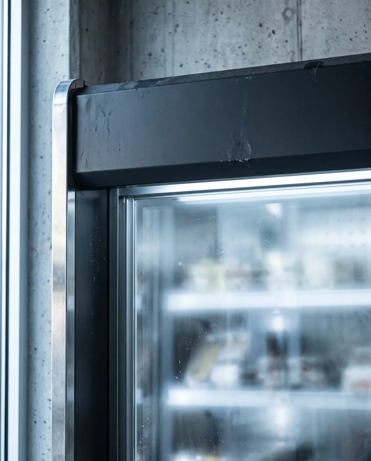 Detail of a matte charcoal retail cabinet, brushed steel trim and backlit glass front against a concrete wall.